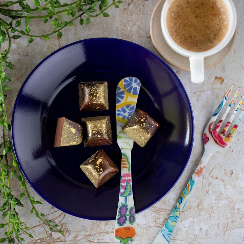 5 Caramel Macchiato chocolates in a blue bowl with a colorful knife, the chocolates angled to show their pyramid shape and brown, gold, and yellow coloring. A cup of coffee is in the corner to evoke the flavor.