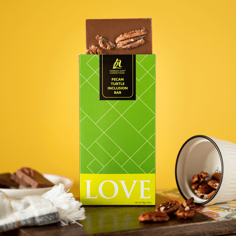 Pecan Turtle Bar (center) in bright green packaging on a yellow background; Square, milk chocolate bar coming out of top of package; Napkin and white plate of chocolate pieces (left background); tilted white bowl with pecans falling out (right). 