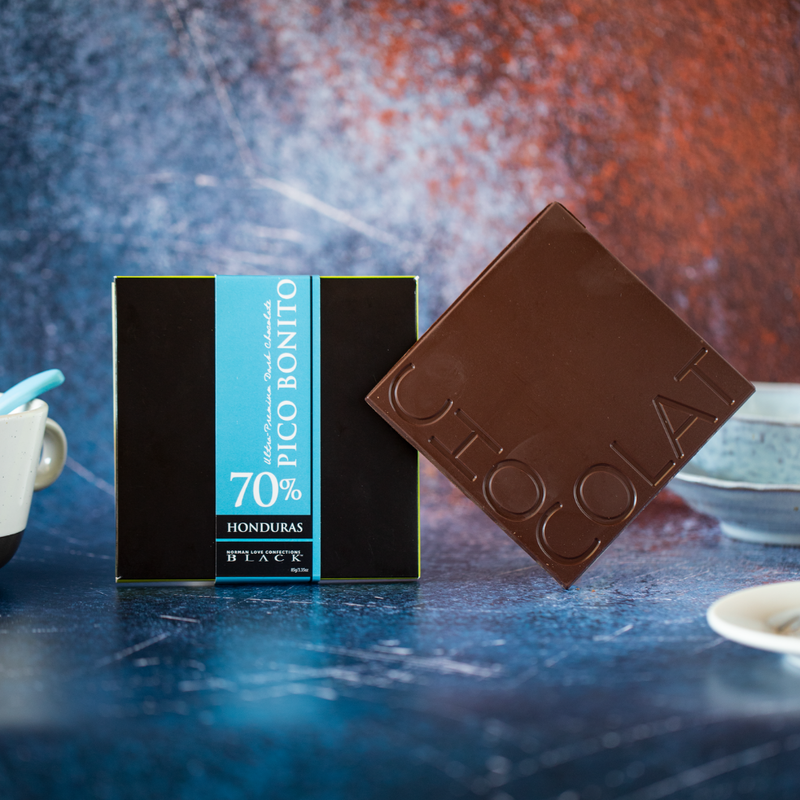 Dark chocolate bar to right of black box with blue label. Blue and brown background. 