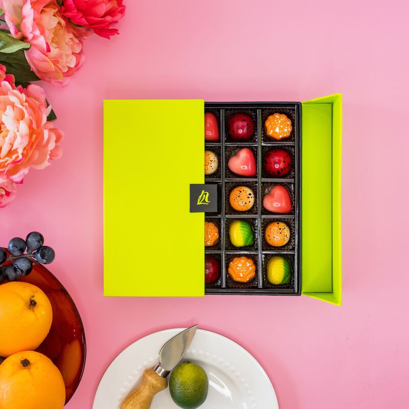 Fruit Favorites Gift Box displays brightly colored chocolates shaped as hearts, seashells, and circles featured in a sleek lime green gift box. The 25 piece box sits on a pink background surrounded with pink flowers, oranges, blueberries and a lime.