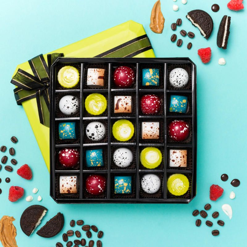 Green 25 Piece Dark Chocolate Box, sitting open atop its lid to display the varieties, colors, and shapes of all 5 flavors. Loose raspberries, coffee beans, cookies & peanut butter are in the periphery to evoke the flavors in the box.
