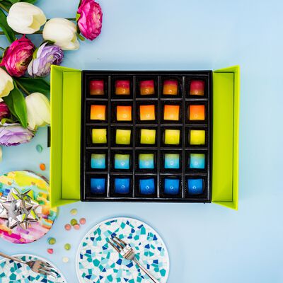 Rainbow colored 25 count box of chocolates on blue background. White and pink flowers, and silver forks (left). 