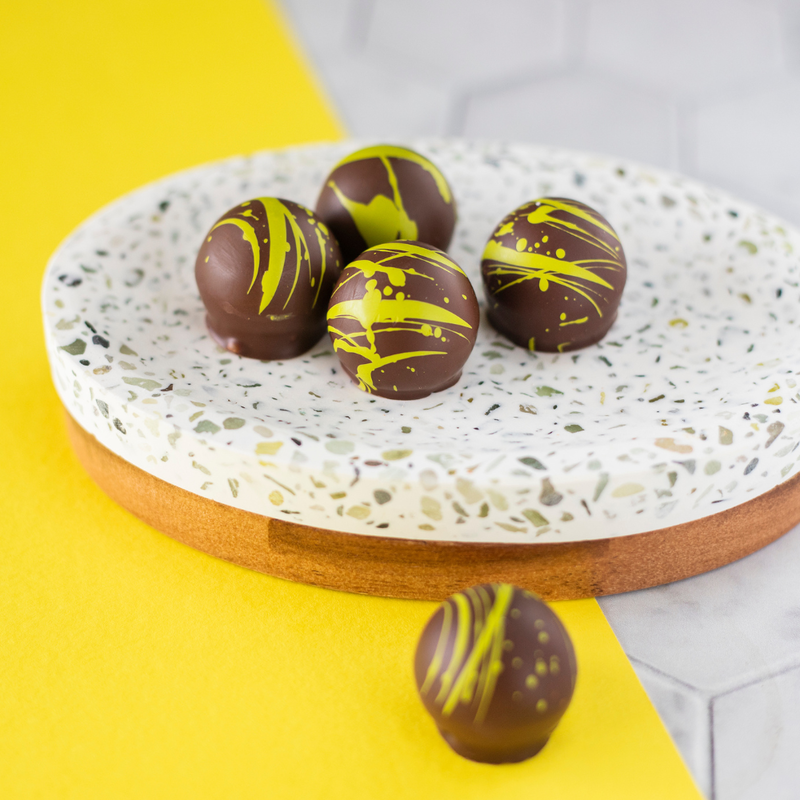 5 dark chocolate cream truffles, 4 on a plate and 1 in front of it, on a piece of yellow paper atop a tile background. Shot at an angle to show the circular chocolates' light green and brown coloration.