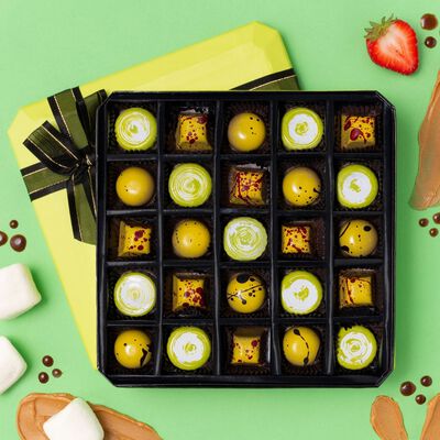 A green 25 Piece Peanut Butter Lovers Gift Box, opened to showcase the shapes, colors, and designs of the included flavors. Marshmallows, strawberries, and smears of chocolate and peanut butter are around the box to evoke the flavors.
