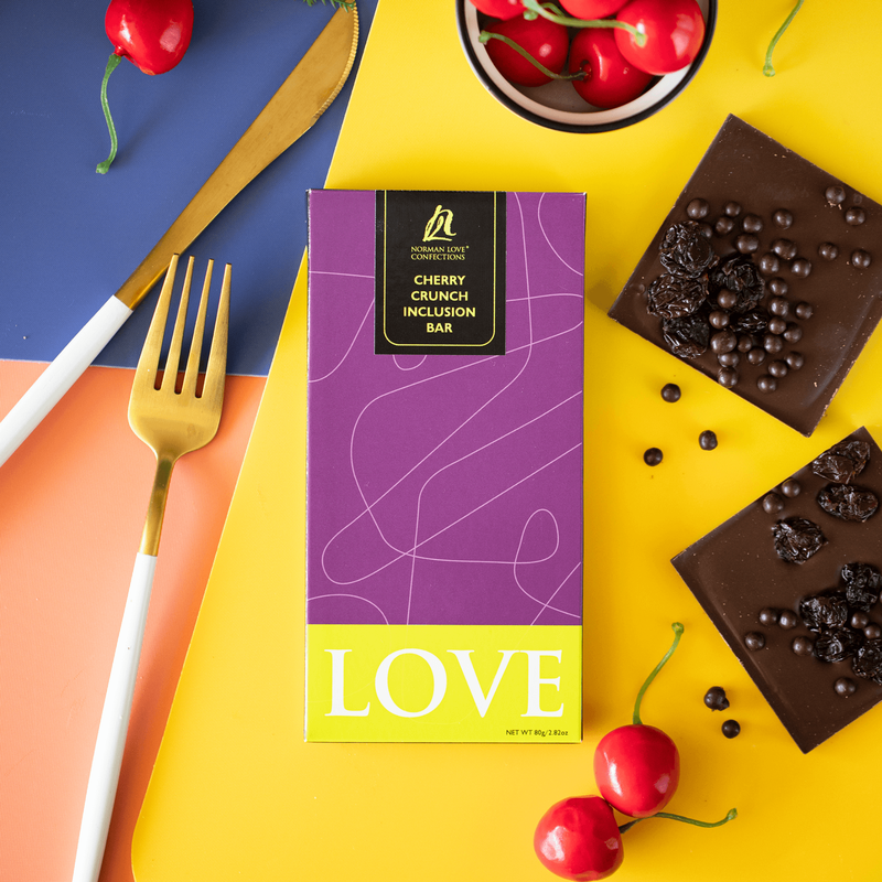 Dark Chocolate Cherry Crunch Bar in purple packaging (center) on blue, orange and yellow background. Gold fork and knife (left) two square dark chocolate pieces (right); cherries (top and bottom).