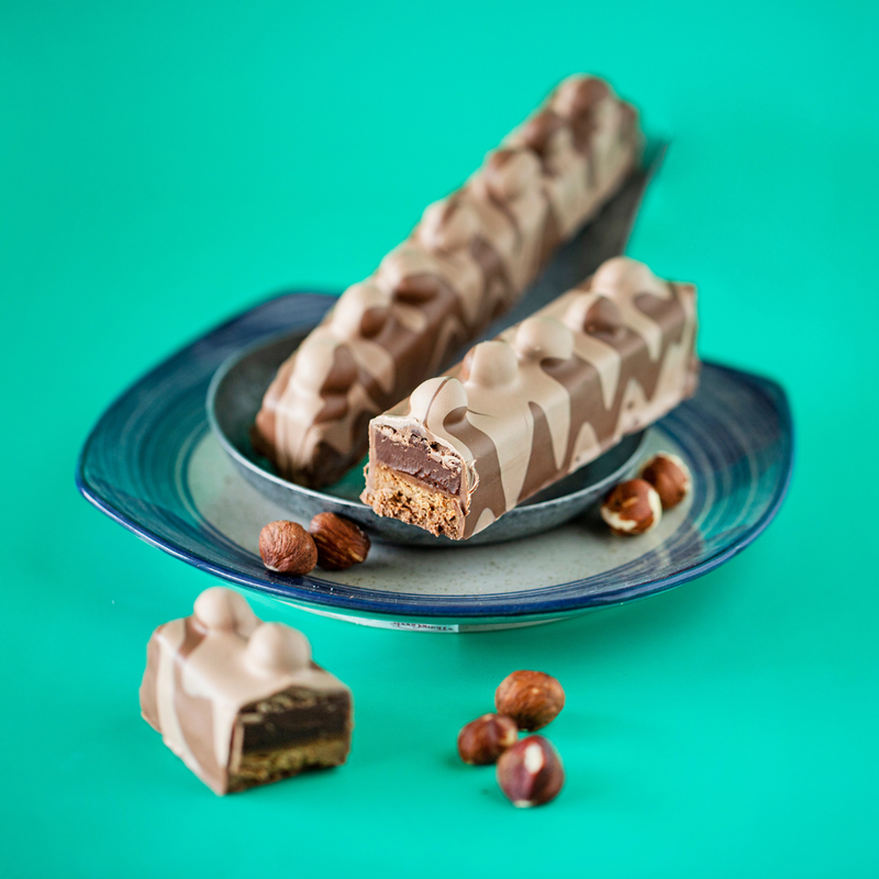 Hazelnut Milk Chocolate bar on dark blue plates on teal background. One whole bar and one broken bar shown on the plate with part of broken bar hazelnuts in foreground.
