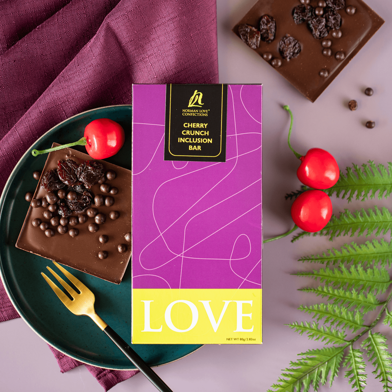 Dark Chocolate Cherry Crunch Bar in purple packaging (center) purple background. Purple napkin and part of square chocolate bar (top); square chocolate bar, gold fork, cherry on dark blue plate (left), cherries and fern leaf (right). 