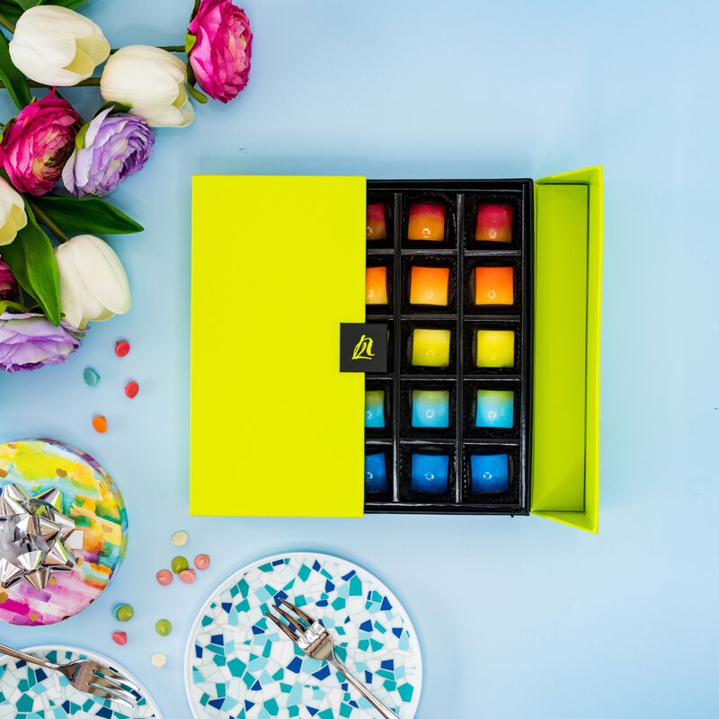 Half closed rainbow colored 25 count box of chocolates on blue background. White and pink flowers, and silver forks (left). 