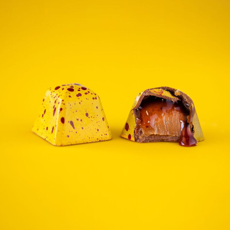 Two Peanut Butter and Jelly chocolates side by side, one intact showcasing it's yellow coloring and red speckles, the other cut in half to display the creamy peanut butter and strawberry jelly interior