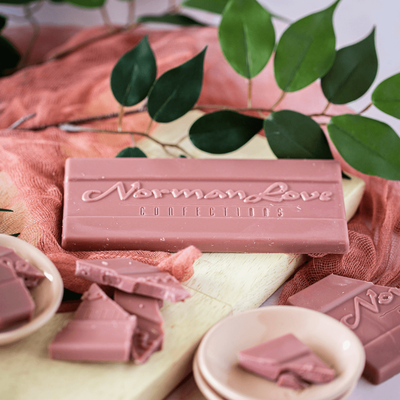 A pink Ruby Cacao Bar on wooden board on pink scarf, with a green leaf in the background and pieces of the pink bar in pink bowls and scattered in the foreground. 