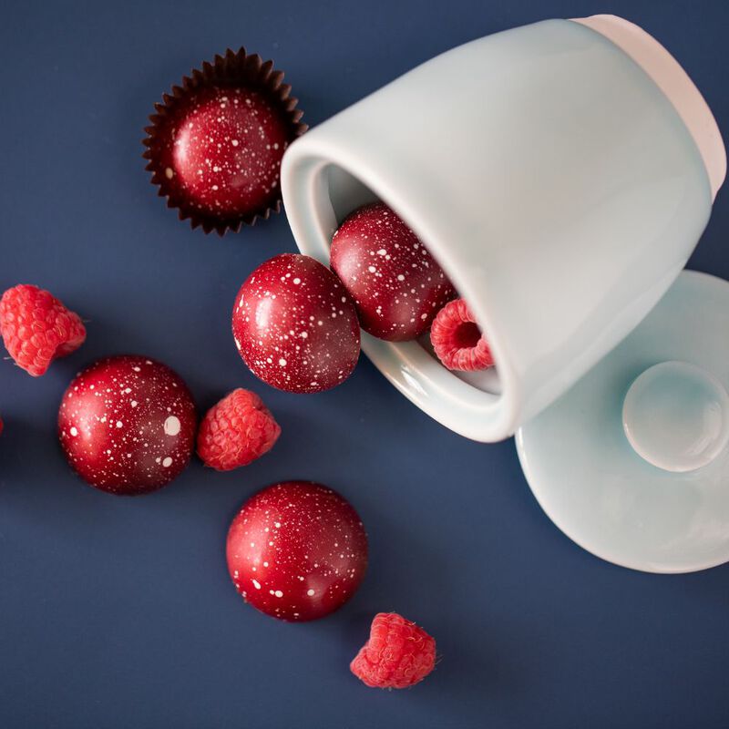 Five raspberry chocolates and individual raspberries spilling out of an overturned coffee cup on a saucer