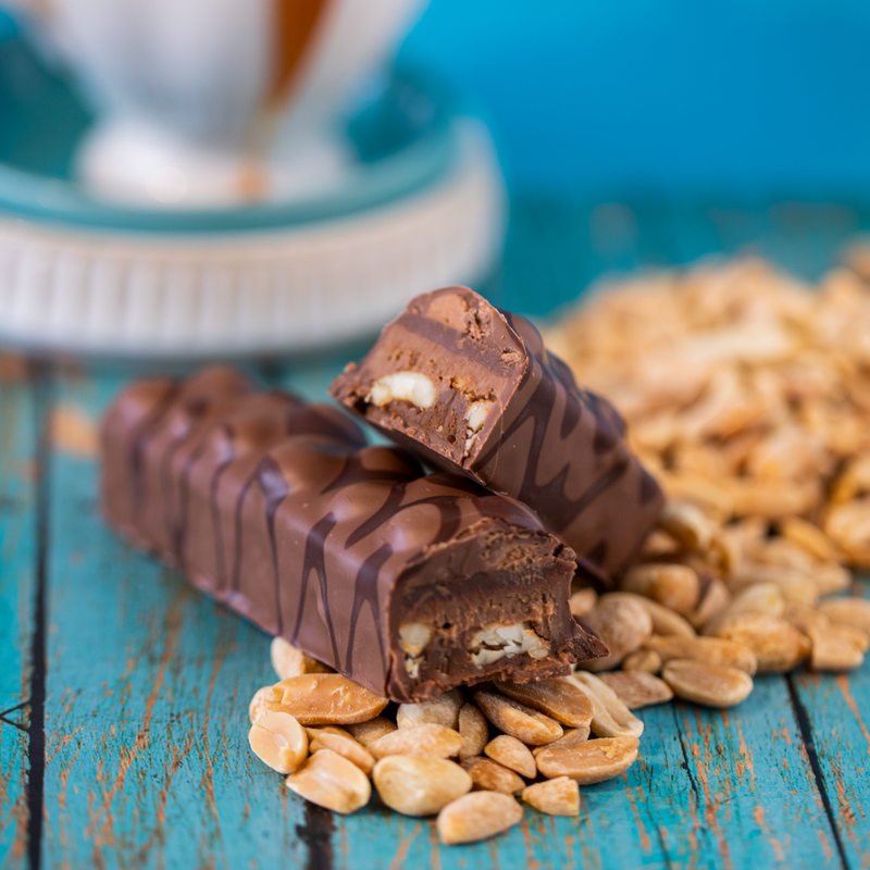 Broken peanut caramel chocolate bar on blue table. Trail of peanuts underneath chocolate bar. White dish (background).