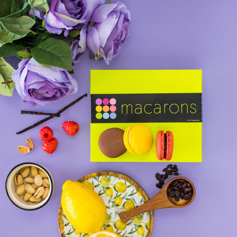 Three French macarons atop a beautiful gift box. Vanilla beans, raspberries, pistachios, lemons and coffee beans surround the gift box to indicate macaron flavors. The purple background and flowers contrast the exquisite lime green box. 