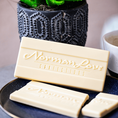 Unwrapped Vegan White Chocolate Bar on black dish. Bar ahs "Norman Love Confections" printed on it. Black planter (background) broken white chocolate bar (foreground). 