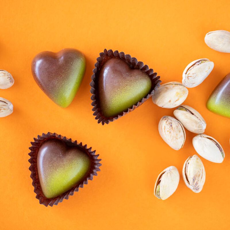 Three Sicilian Pistachio Chocolate hearts, two in candy wrappers, one without a wrapper, on an orange background with pistachios in shells on the right side 