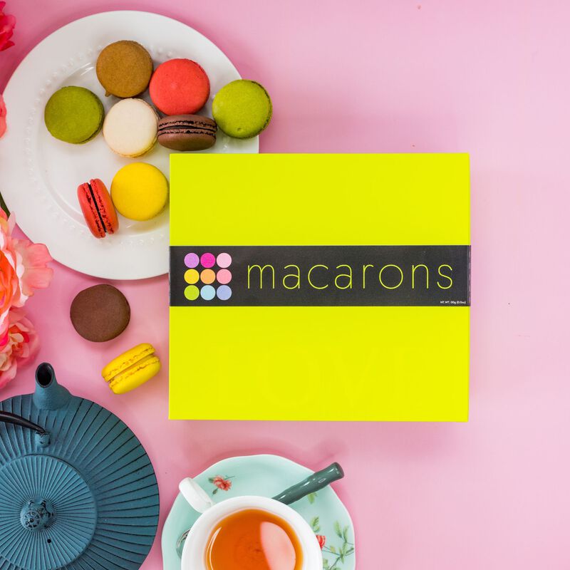 An overhead shot of a stunning Macaron Gift Box on a pink background. 10 macarons of various colors and flavors are shown on a white plate next to the box. A blue tea kettle and cup of tea are shown below the box. 
