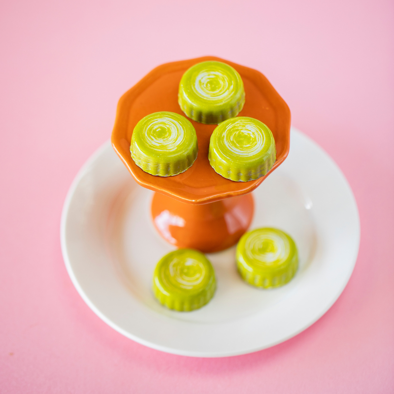 5 Peanut Butter Cup Chocolates, 3 sitting atop an orange pedestal which sits on a white plate with the other 2 pieces. Shot at an angle to show the scalloped edge & green color of the chocolate.