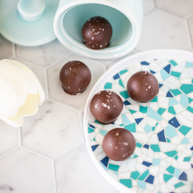 5 sea salt caramel truffles sitting on a blue plate on a white tile surface. Shot at an angle to show the sea salt sprinkled atop each piece with a white rose in the periphery.