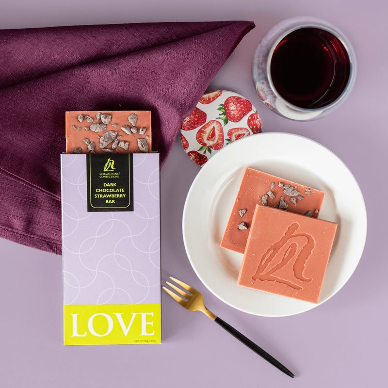 Dark chocolate strawberry bar in light purple packaging (left); Unwrapped pink bar coming out of package & on white dish( right), dark purple napkin (top right) strawberry lid & cup of tea (top right), gold fork (bottom) on light purple background.