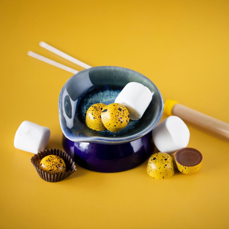 5 Peanut Butter Marshmallow chocolates arranged on and around a blue ceramic dish. The bonbons are round and colored yellow dotted with black. Marshmallows and a skewer are in the background to evoke the flavor.