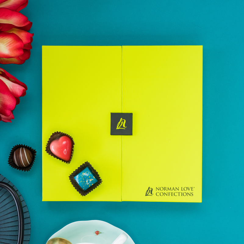 One pink heart-shaped and one blue square chocolate atop a closed 25- Piece Signature Gift box. To the side of the box is a dark brown chocolate along with red roses. The stylish gift box is accented by the dark teal background. 