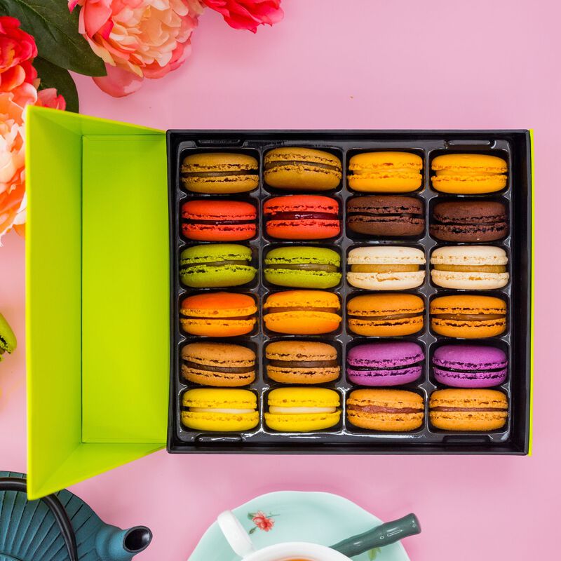 A stunning Macaron Gift Box on a pink background. The gift box displays 24 brightly colored macarons in 12  flavors: PB&J, Lemon, Salted Caramel, Passionfruit, Cassis, Coffee, Pistachio, Hazelnut, Dark Chocolate, Raspberry, Mango Lime, and Vanilla.