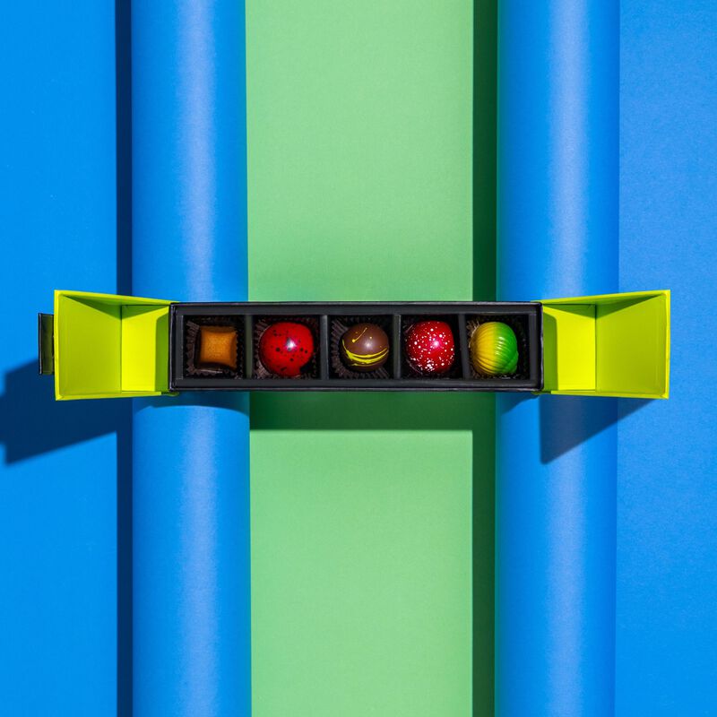 A five piece signature gift box on a blue and green background. Gift box is open to show five pieces from the signature chocolate collection: Strawberry, Key Lime, Tahitian Caramel, Raspberry, Dark Chocolate Cream Truffle.