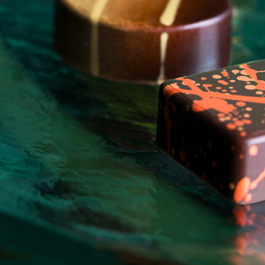 Two chocolate truffles, one with yellow and one with orange decoration on green background.